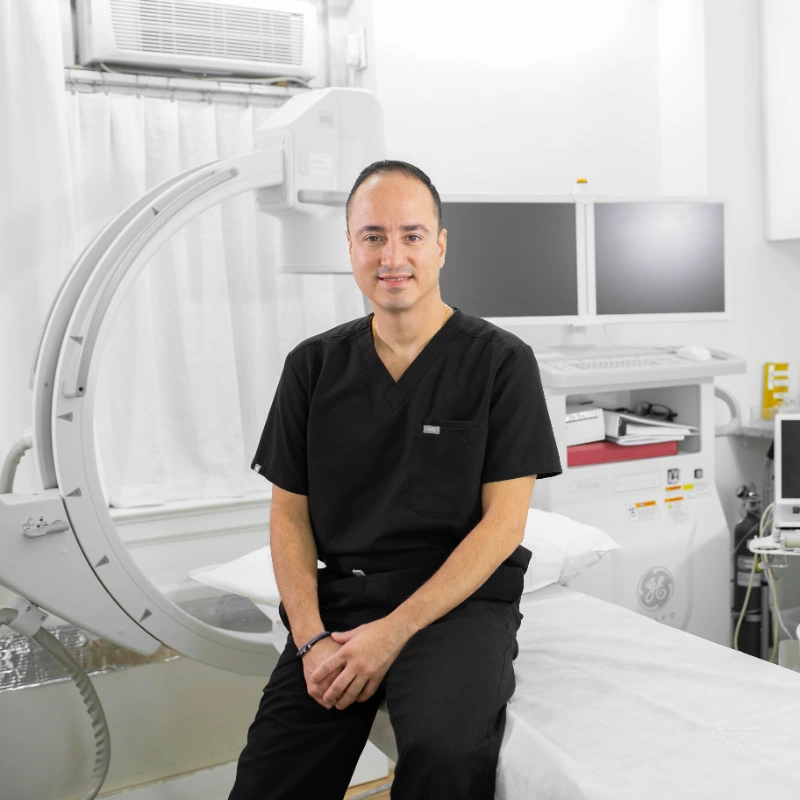 Montoya IMG_lifestyle_optimized Dr. Montoya positioned in a bright, modern clinic room equipped with advanced medical machinery, including a large C-arm imaging system to his left.