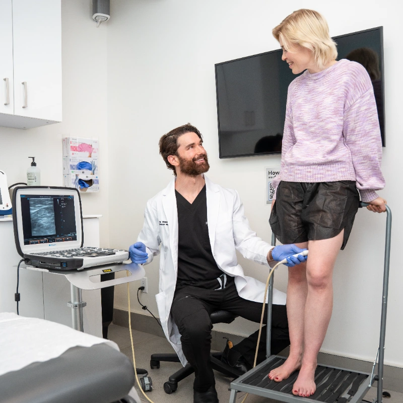 Dr. Shaun Cole, MD performs a Duplex Ultrasound examination of a patient's leg at the Vein Treatment Clinic using a portable Ultrasound Machine to assess for Varicose Veins, Spider Veins, Venous Reflux, and Chronic Venous Insufficiency. The patient stands on a scale platform while Dr. Cole operates the handheld Ultrasound Probe with blue gloves in this modern clinical setting.